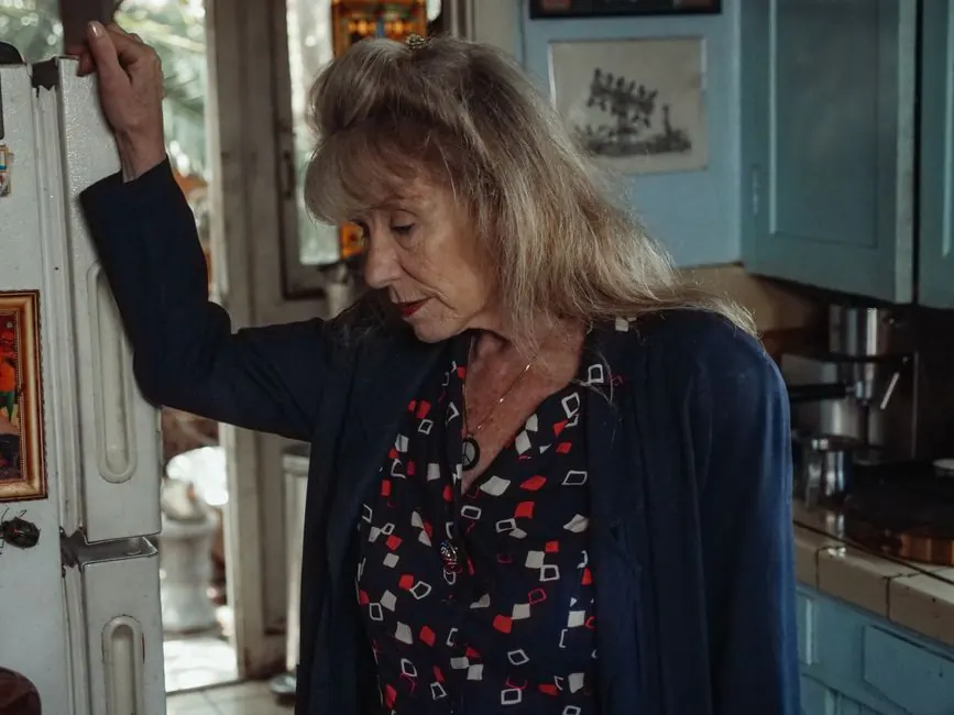 A thoughtful senior woman leans on a refrigerator, evoking emotion and nostalgia in a cozy kitchen setting.